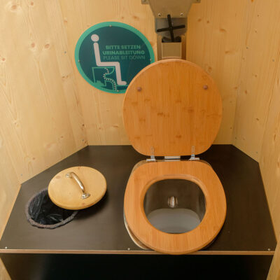 Innenansicht des Humero Rollin’ Toilettenanhängers mit Holz-Toilettensitz, geöffnetem Deckel, Mülleimer, Urinableitungssystem, separatem Einstreubehälter und Sticker an Holzwand.
