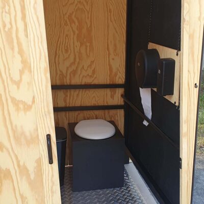 Libre Event 13 Ansicht durch Tür auf barrierefreie Trockentoilette mit weißer Klobrille und geschlossenem Deckel auf Metallboden, Papierhalter und Desinfektionsspender an Holzplatte für hygienische Nutzung.