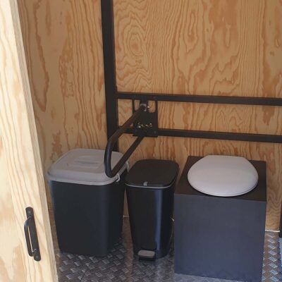 Libre Event 11 Ansicht durch Tür auf barrierefreie Trockentoilette mit weißer Klobrille und hochgeklapptem Deckel, schwarzem Haltegriff, Mülleimer und Eimer mit Stroh zum Einstreuen auf Metallboden für komfortable und hygienische Nutzung.