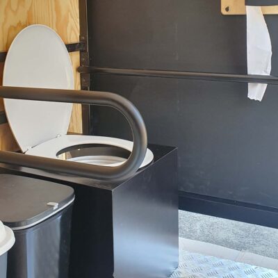 Libre Event 12 Offene barrierefreie Trockentoilette mit weißer Klobrille und hochgeklapptem Deckel, schwarzem Haltegriff und Mülleimer auf Metallboden für komfortable und hygienische Nutzung.