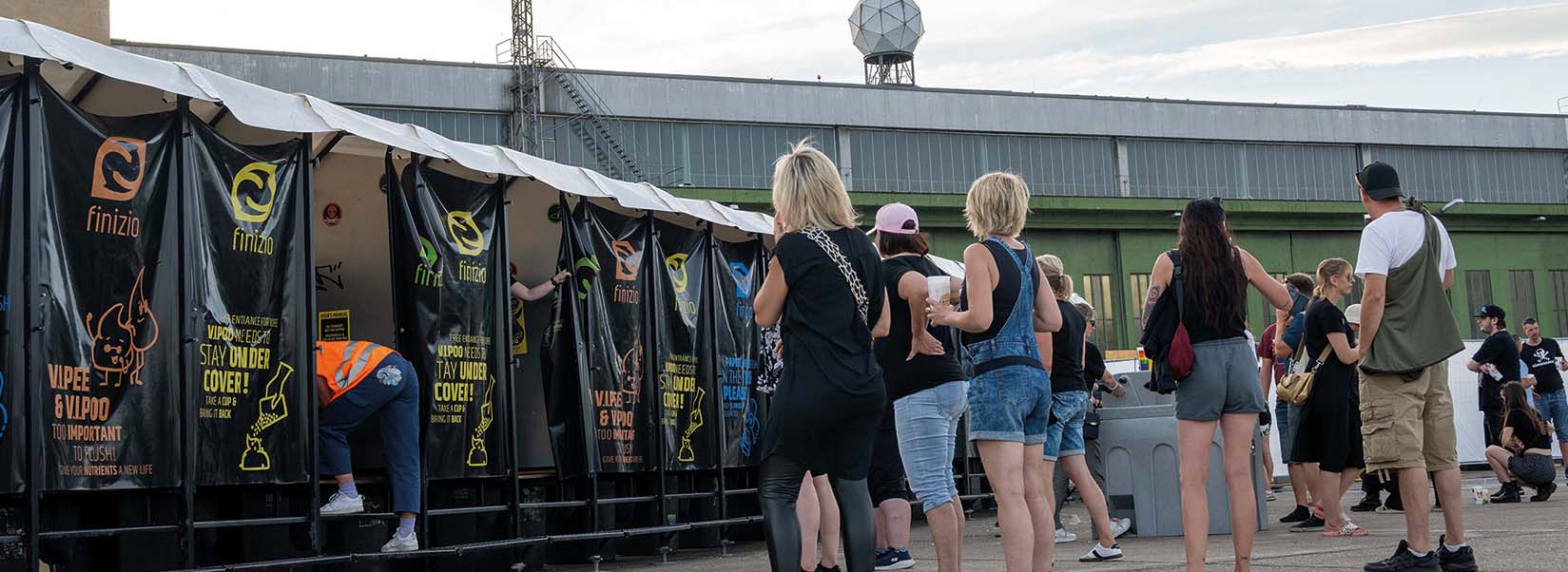 2024_©Finzio_GmbH_Drytoilets_and_Recycling_Trockentoiletten_Trenntoiletten_Festivaltoilette_Humussphaere_Headerbild_Tempelhoferfeld Mehrere Personen stehen vor mobilen Finizio-Trockentoiletten in einer Schlange, während eine Person in Warnweste eine Kabine sauber macht.