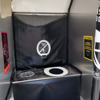 Innenansicht der Humusspähre Trockentoilette mit schwarzer Rückwandplane, weißer, aufgeklebter Toilettenbrille, Mülleimer und Bedienhinweisen.