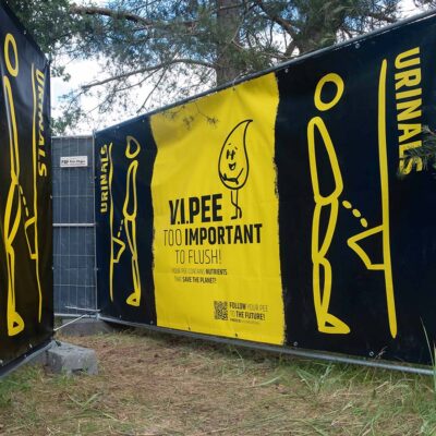 Peeter 2 Schwarze und gelbe Sichtschutzbanner mit Urinal-Symbolen und Hinweis V.I.Pee an Bauzaun im Außenbereich.
