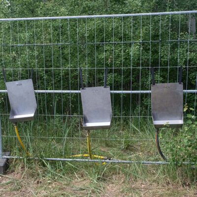 Peeter 3 Drei Peeter Herrenurinale aus Edelstahl an Bauzaun montiert mit gelben und schwarzen Schläuchen im Außenbereich.
