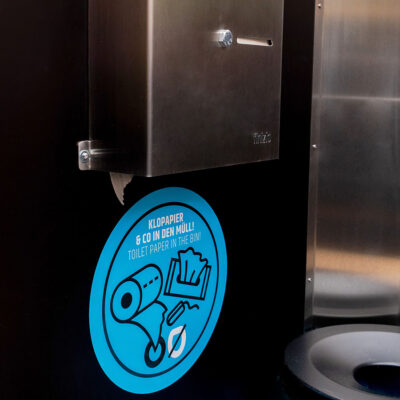 Libre Light 9 Toilettenpapierspender aus Edelstahl in der Libre Trockentoilette mit blauem Hinweisschild.