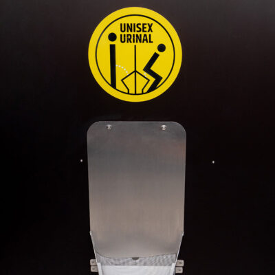 Libre Light 8 Edelstahl-Unisexurinal in der Libre Trockentoilette vor schwarzer Wand mit gelbem Hinweisschild: Unisex Urinal