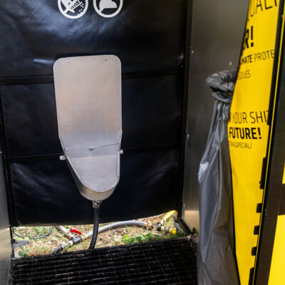 Unipee 1 Innenansicht des Unisexurinals mit Edelstahlbecken, schwarzer Rückwand und Müllbeutelhalterung neben gelber Türplane.