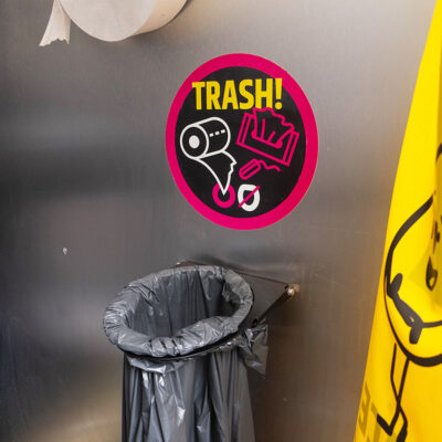 Unipee 2 Innenansicht des Unisexurinals Unipee mit rundem Trash-Hinweis-Sticker für Papierabfall und schwarzem Müllbeutel an Metallwand.