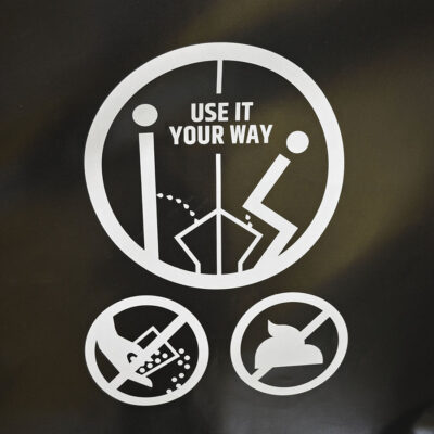 Weißes Piktogramm mit Text „Use it your way“ und Symbolen zur Nutzung des Unisexurinals Unipee auf schwarzem Hintergrund an Rückwandplane.