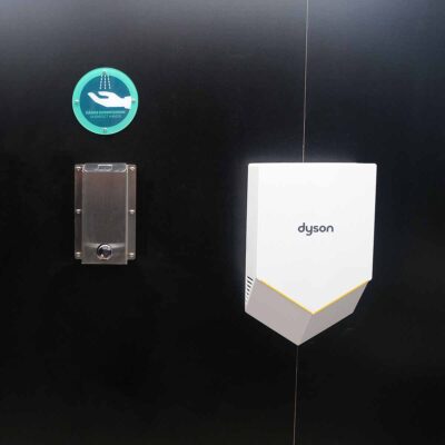 Libre DIN 9 Detailansicht des Dyson Händetrockners und Edelstahl-Desinfektionspender in der Libre-DIN Kabine mit Hinweisaufkleber.