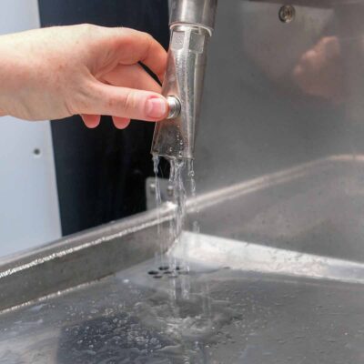 Libre DIN 14 Detailansicht einer Hand, die den Wasserhahn der Libre-DIN Trockentoilette betätigt, mit fließendem Wasser.