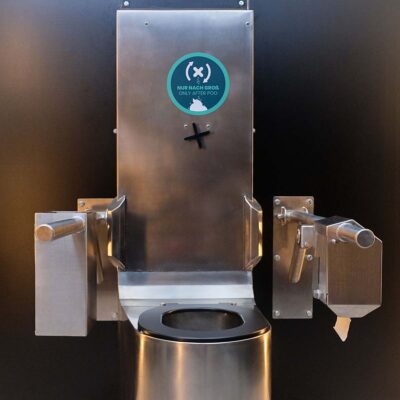 Libre DIN 5 Frontansicht der rollstuhlgerechten Libre-DIN Trockentoilette aus Edelstahl mit Klappstützgriffen und Hinweisaufkleber.