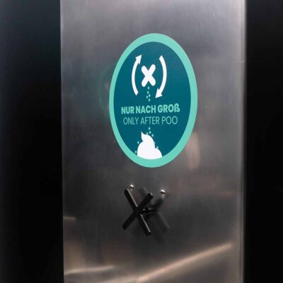 Libre DIN 16 Detailansicht der Edelstahlfläche des Streuspenders Spencer mit Hinweisschild mit Text „Nur nach Groß – / Only after poo“ und Symbol für Einstreu-Nutzung nach dem Toilettengang.