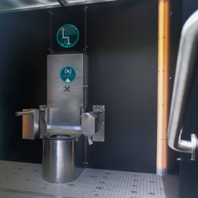Libre DIN 2 Innenansicht Libre DIN barrierefreie, geschlechterneutrale Trockentoilette mit Edelstahltoilette und Klappstützgriffen an schwarzer Wand.