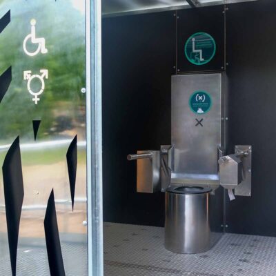 Libre DIN 1 Innenansicht Libre DIN barrierefreie, geschlechterneutrale Trockentoilette mit Edelstahltoilette und Klappstützgriffen an schwarzer Wand.