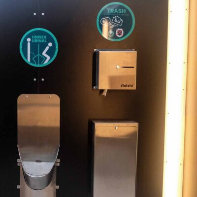 Libre DIN 7 Innenansicht Humero Trockentoilette mit Unisexurinal aus Edelstahl, Papierhandtuchspender und Abfallbehälter an schwarzer Wand.
