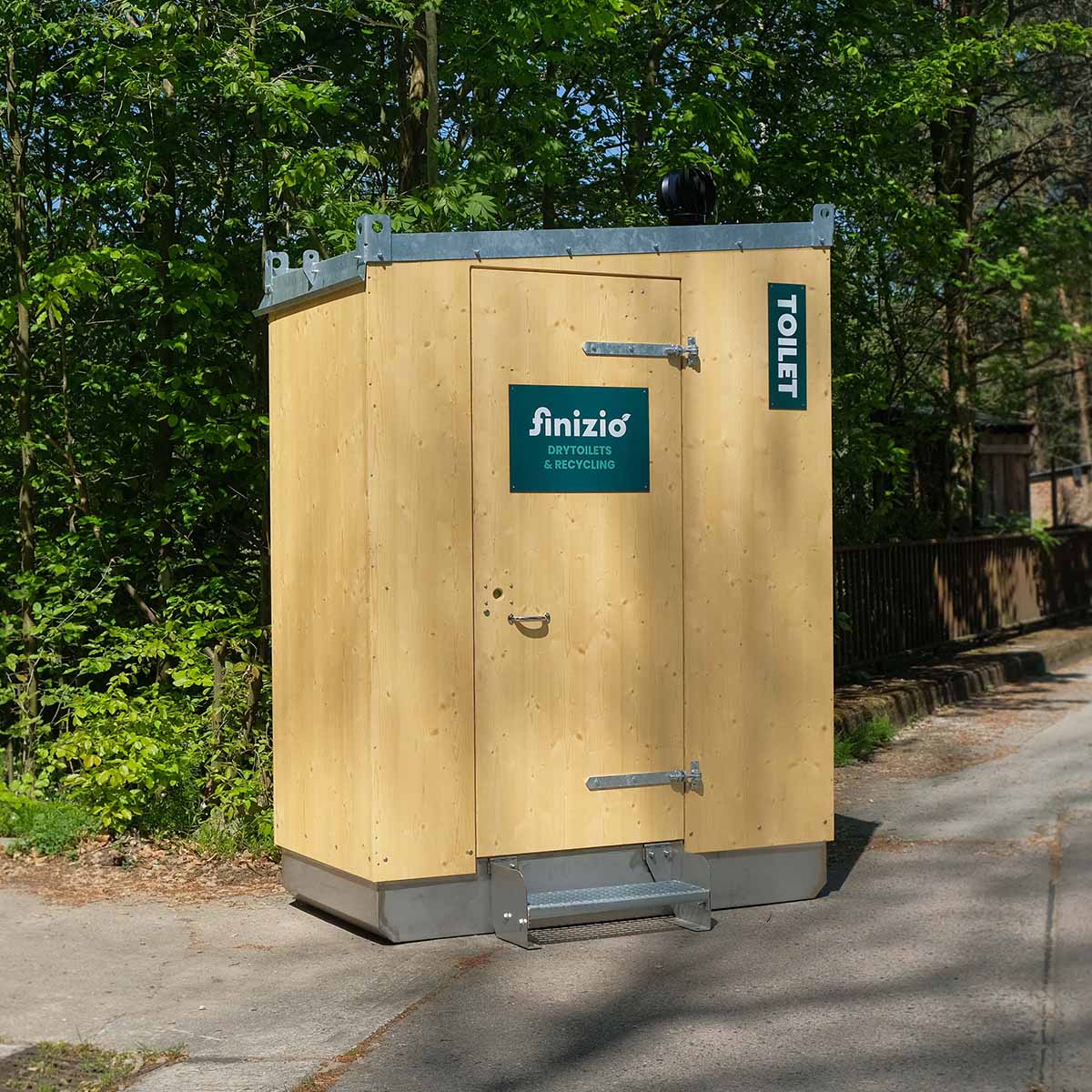 Humero Trockentoilette mit Holzverkleidung und geschlossener Holztür vor grünem Wald.