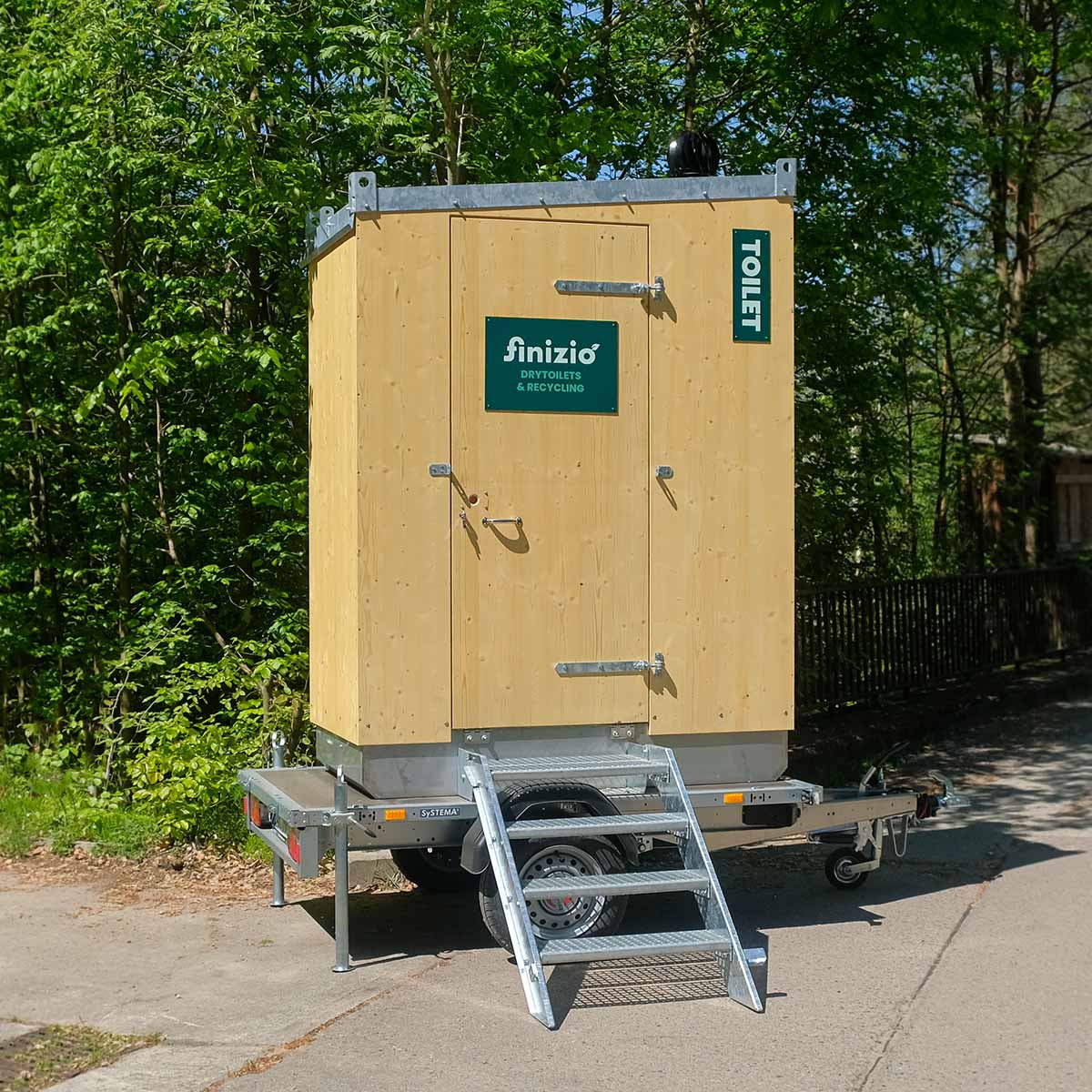 Kontakt 11 Humero Rollin’ Trockentoilette auf Trailer mit Holzverkleidung und Metalltreppe vor grünem Hintergrund