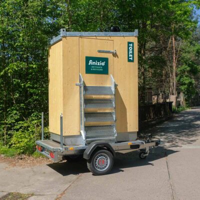 Humero Rollin’ Trockentoilette auf Trailer mit Holzverkleidung und hochgeklappter Metalltreppe vor grünen Päumen.