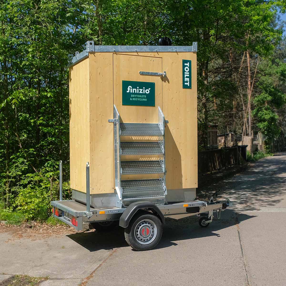 Humero Rollin’ Trockentoilette auf Trailer mit Holzverkleidung und hochgeklappter Metalltreppe vor grünen Päumen.