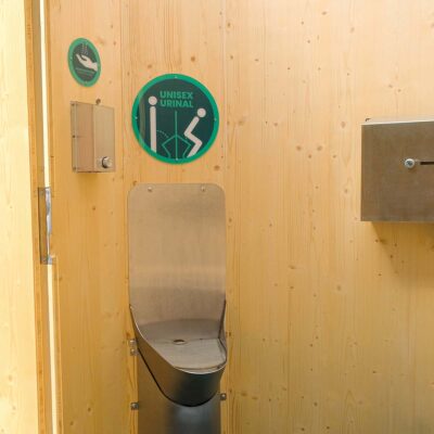 Unisexurinal, Desinfektionsmittelspender und Toilettenpapierspender aus Edelstahl sowie zwei Hinweisschilder in der Humero Rollin’-Toilettenkabine.