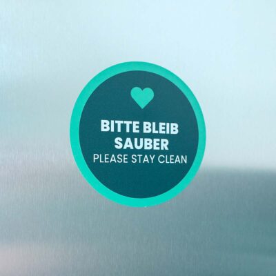 Runder Sticker mit weißem Text „Bitte bleib sauber / Please stay clean“, darüber grünes Herz-Symbol.