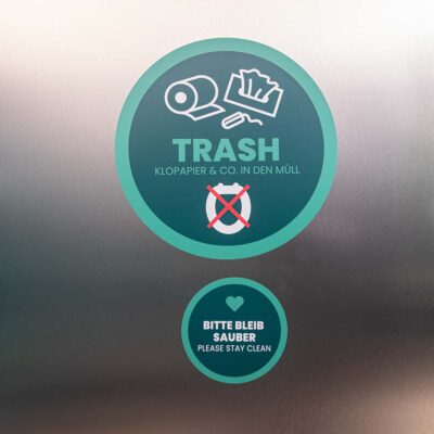 Zwei Sticker auf Metallinnenwand der Kabine: rund, petrolfarben, oben mit weißem Piktogramm und Text „Trash / Klopapier & Co in den Müll“, unten kleiner rund mit weißem Text „Bitte bleib sauber / Please stay clean“.