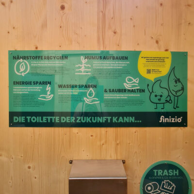 Neues Design: Rechteckiges grünes Informationsschild mit Text „Die Toilette der Zukunft kann…“ und Icons zu Recycling, Humusaufbau, Energie- und Wassersparen sowie sauber halten an Holzwand.