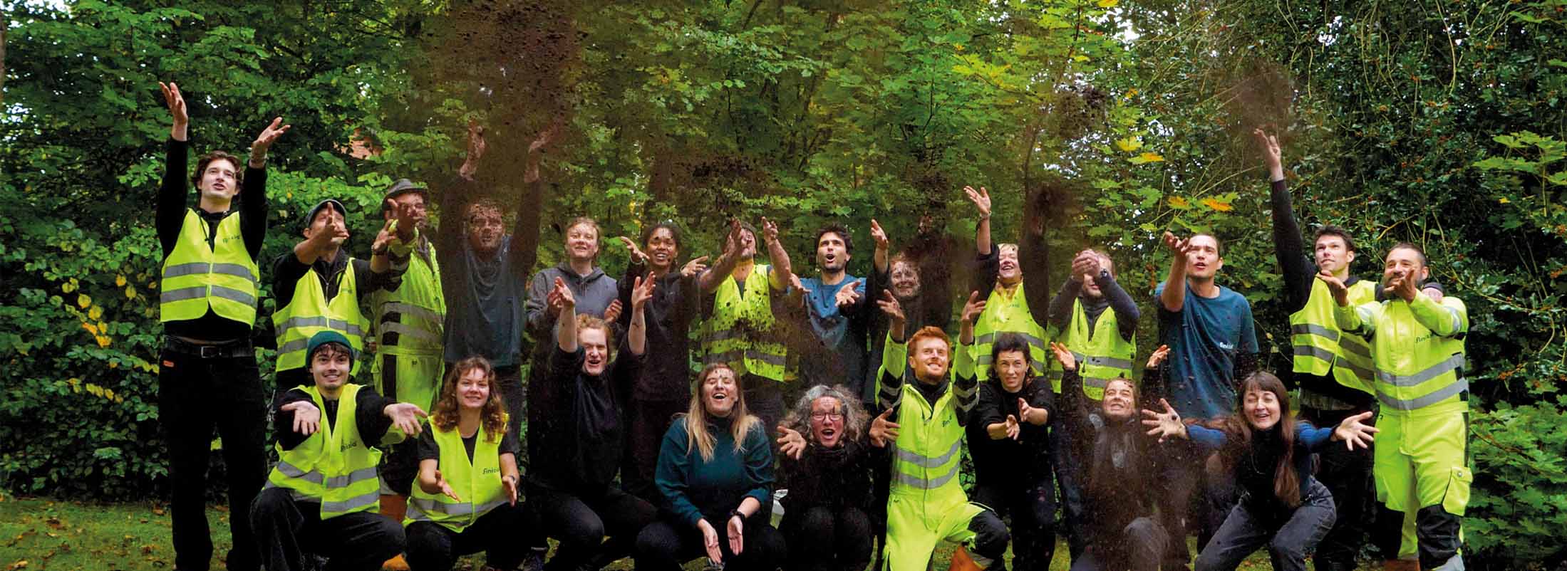 Personen in Warnwesten werfen Humusdünger aus Trockentoiletteninhalten in die Luft vor einem grünen Wald.