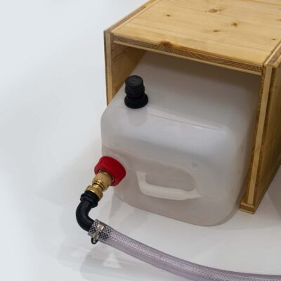 Nahaufnahme des 10-Liter-Urintanks aus Kunststoff mit angeschlossenem Ablaufschlauch und Messingkupplung im Holz-Beistelltisch.
