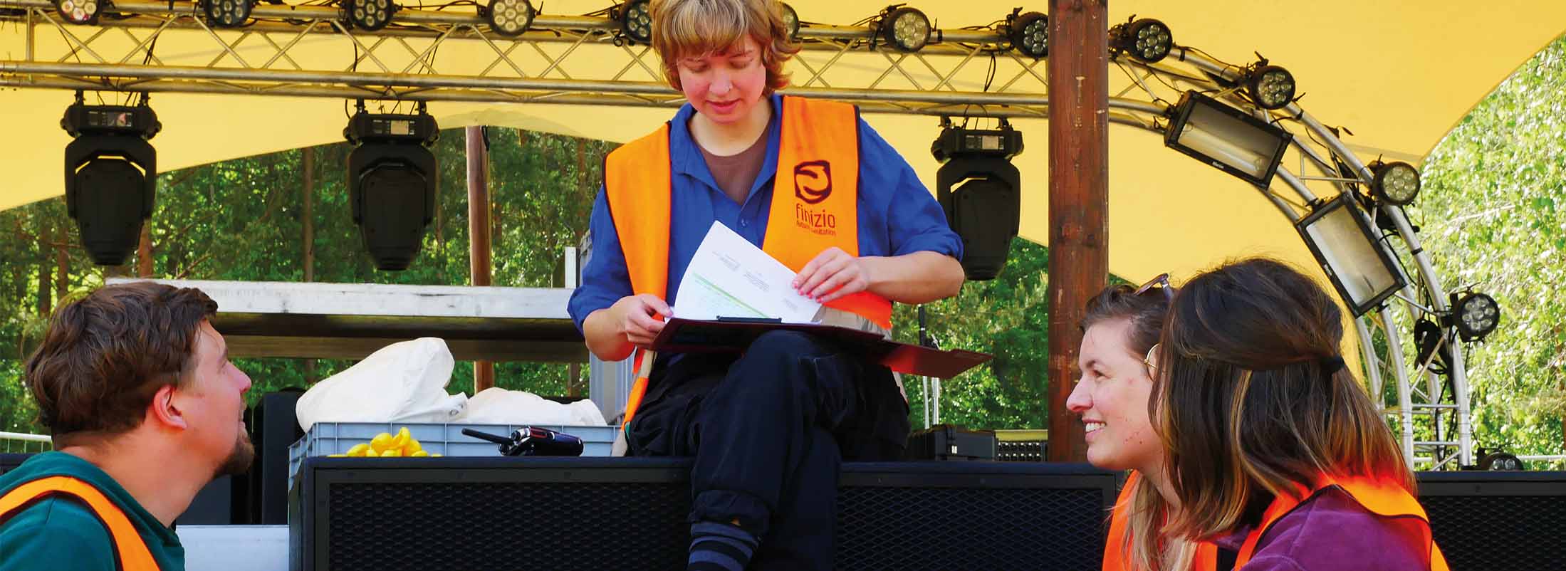 Vier Personen in Westen sitzen auf einem Festival, eine Person hält ein Klemmbrett.