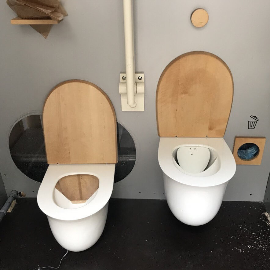 Zwei Trenntoiletten mit Holzdeckel und Keramikschüssel nebeneinander in einem grauen Ausstellungsraum.