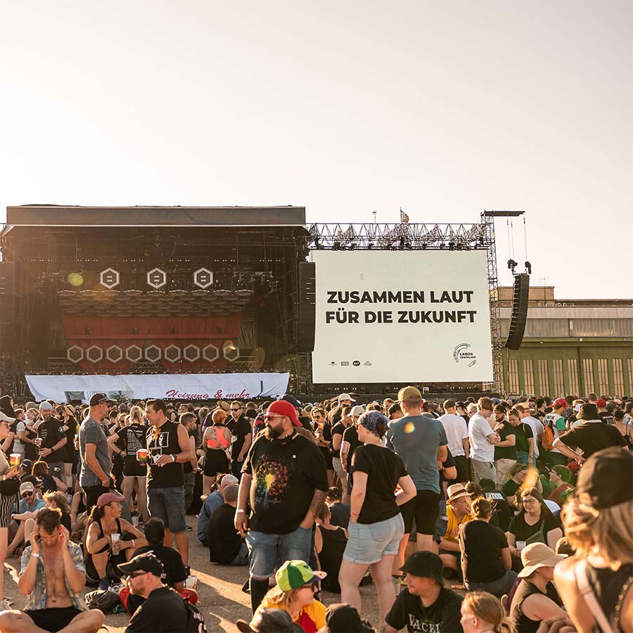 C2C_7355_COPYRIGHT PAUL GÄRTNER - FOTOGRAF-330 Viele wartende und sitzende Konzertbesuchende auf einem Outdoor Konzert, im Hintergurnd eine große Bühne, auf deren Screen "Zusammen laut für die Zukunft" steht.